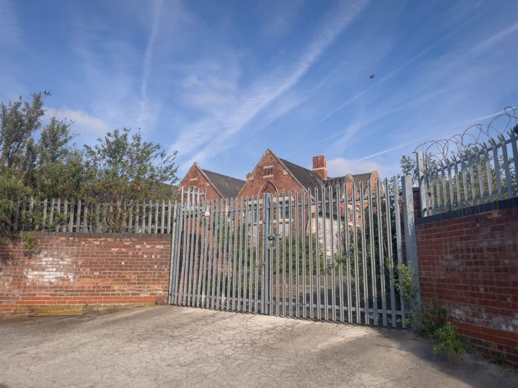 A tall metal gate with barbed wire surrounds an old, overgrown red brick building under a blue sky with wispy clouds. Vegetation grows through the fence, and part of the building is obscured.