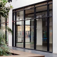 A modern building with large glass windows and doors, reflecting nearby structures. Indoor plants and a wooden bench are visible in the foreground, creating a clean, contemporary outdoor space.