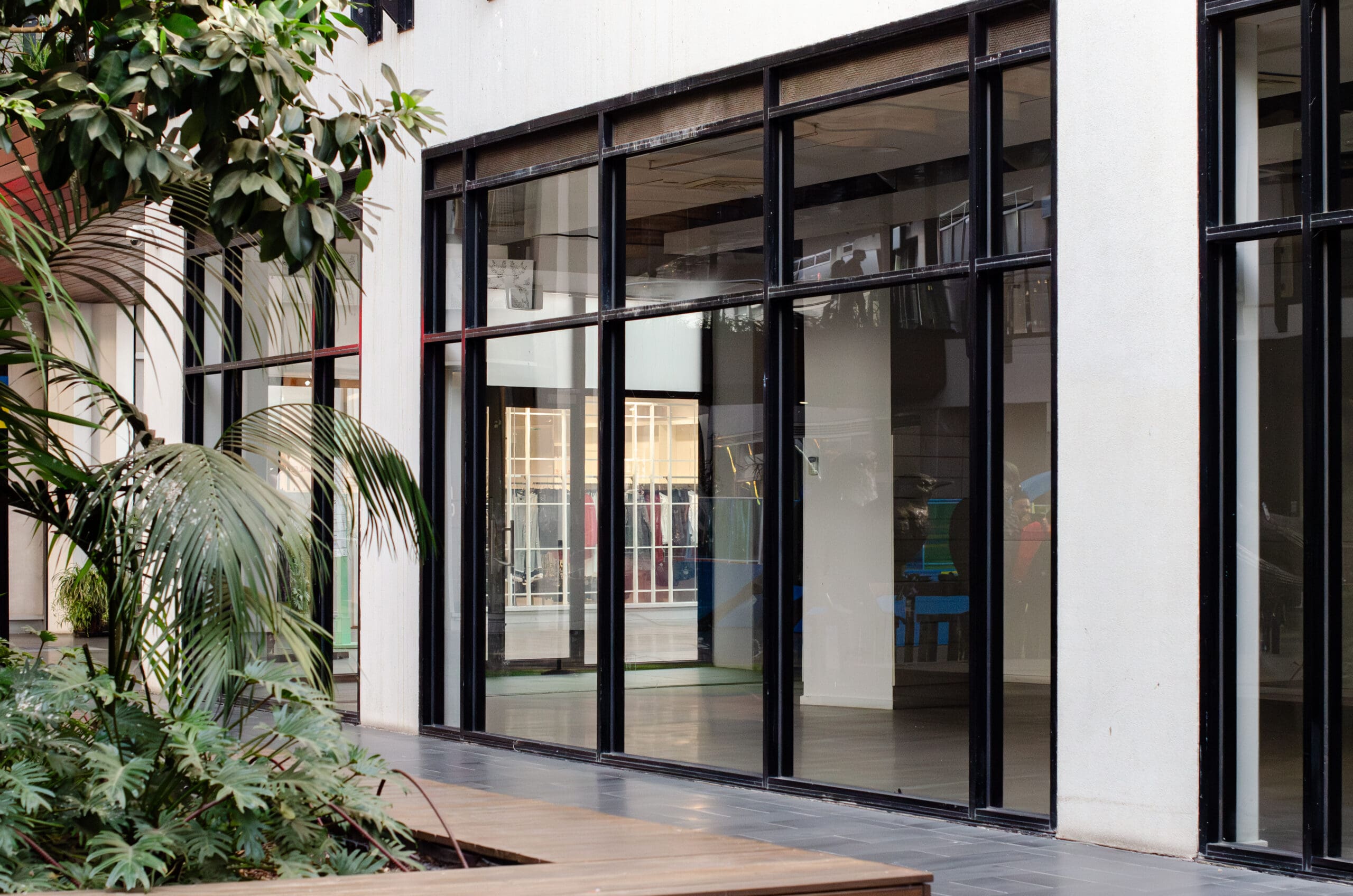 A modern building with large glass windows and doors, reflecting nearby structures. Indoor plants and a wooden bench are visible in the foreground, creating a clean, contemporary outdoor space.