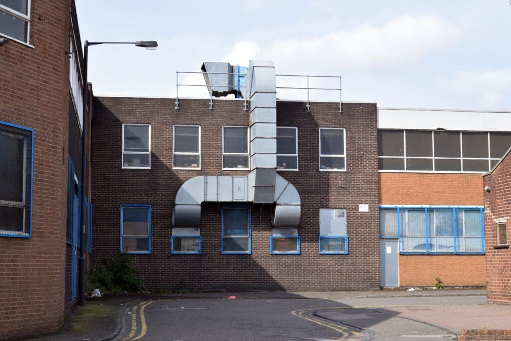 A brown brick industrial building with large metal ventilation ducts on the exterior and several windows framed in blue; the street in front is empty with yellow lines and a bit of vegetation at the building's base.