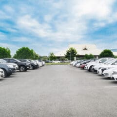 A large outdoor parking lot with rows of parked cars on both sides, mostly white and silver, under a bright blue sky with scattered clouds. Green trees and a building are visible in the background.