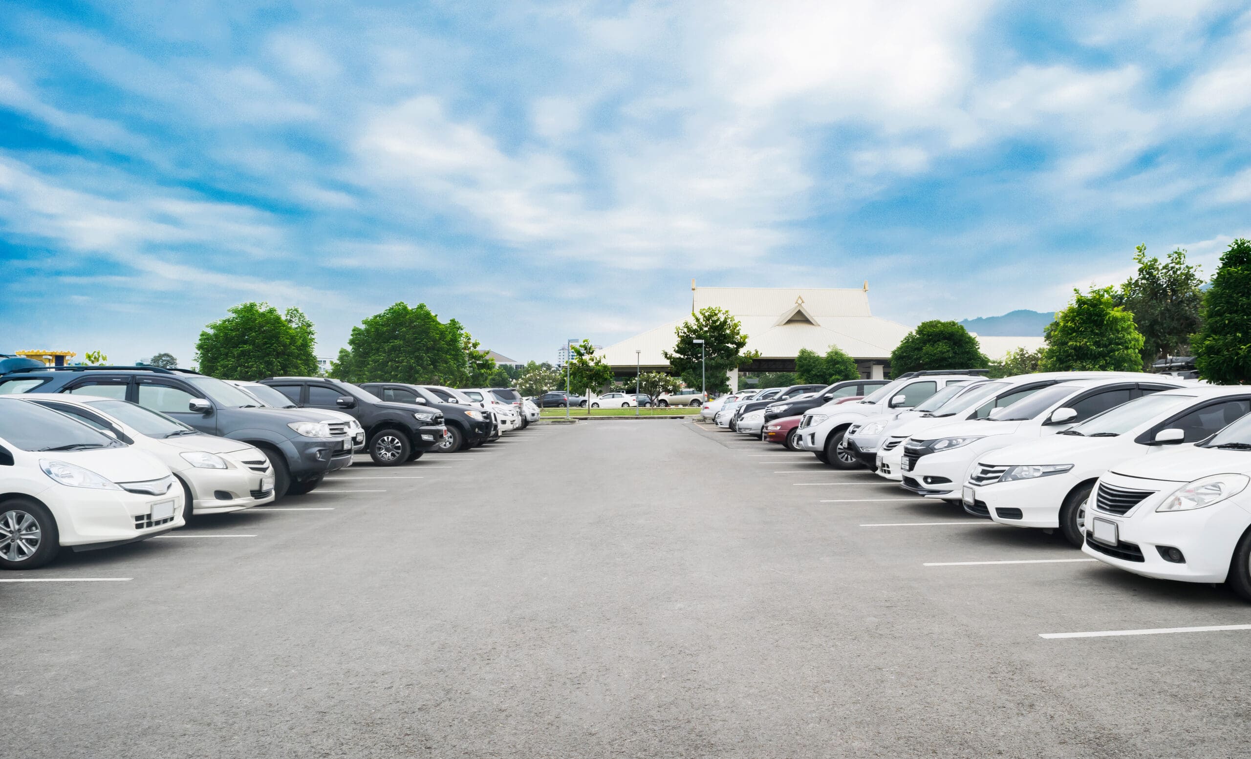A large outdoor parking lot with rows of parked cars on both sides, mostly white and silver, under a bright blue sky with scattered clouds. Green trees and a building are visible in the background.