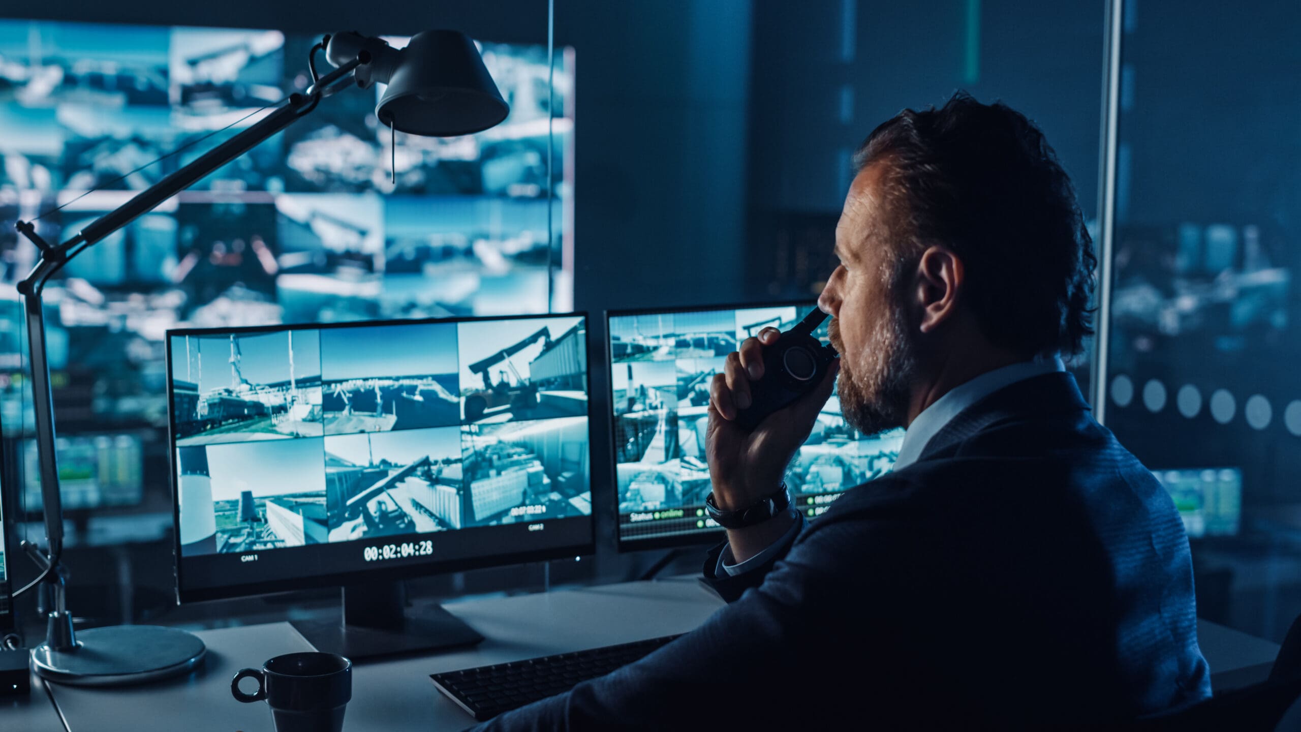 A man in a suit sits at a desk, monitoring multiple surveillance camera feeds on computer screens in a dimly lit control room, speaking into a walkie-talkie.