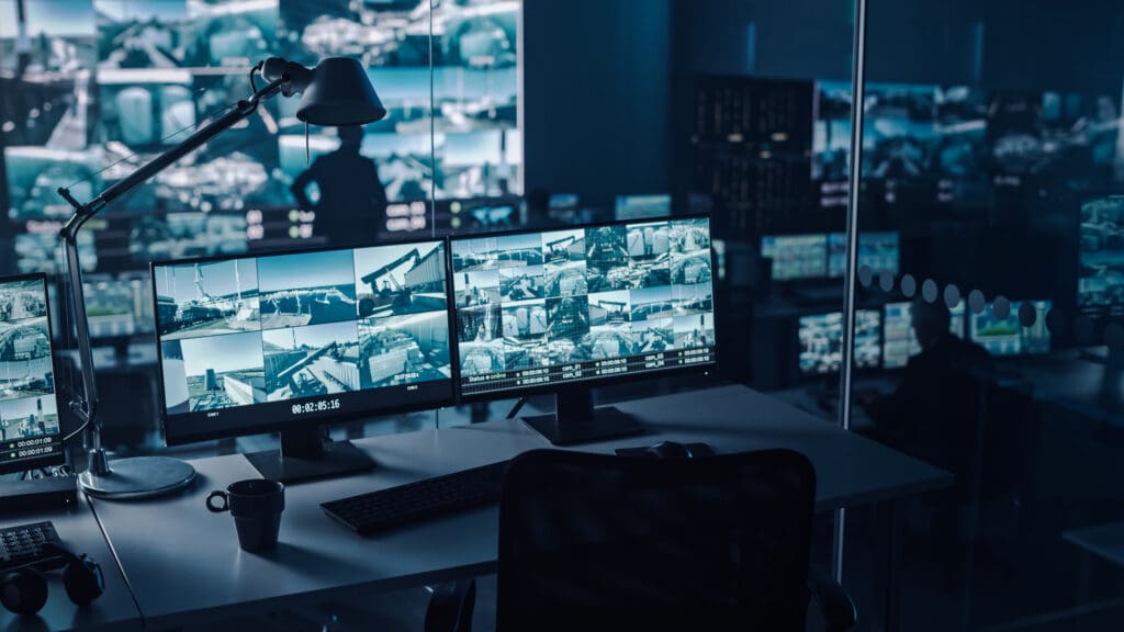 A modern security control room with multiple monitors displaying surveillance camera footage, a keyboard, a lamp, and a coffee cup on the desk. Reflections and blurred figures are visible in the background.