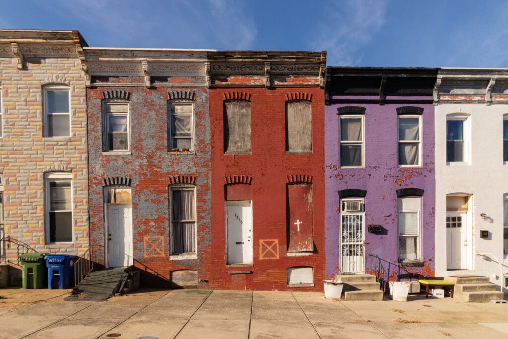 Three row houses are shown: one with tan stone siding, one with worn red brick and boarded windows, and one with faded purple paint. All appear weathered, and the red house’s windows and door are covered with boards.