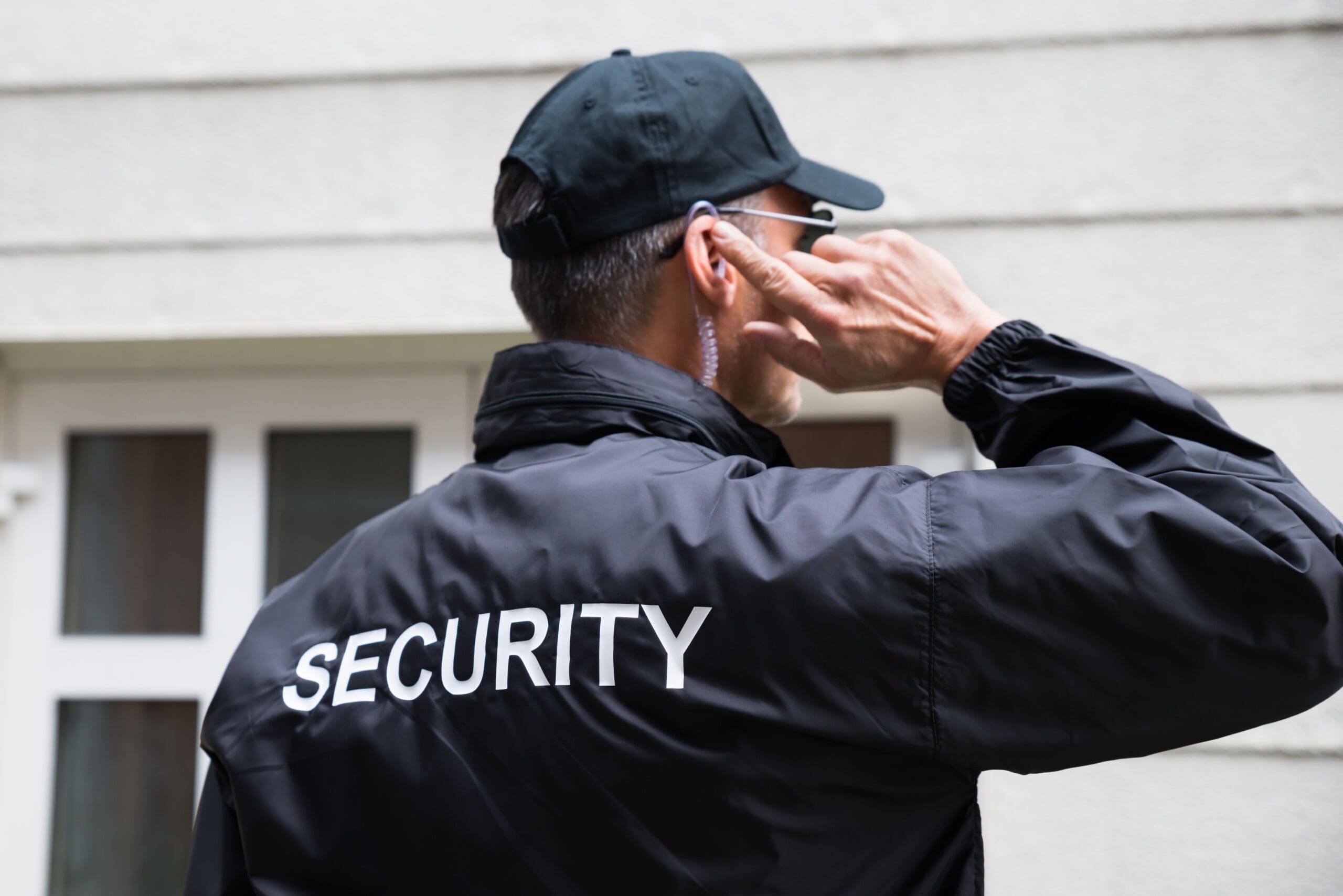 A security guard wearing a black jacket labeled "SECURITY" and a black cap stands outside a building, holding an earpiece with one hand while facing away from the camera.