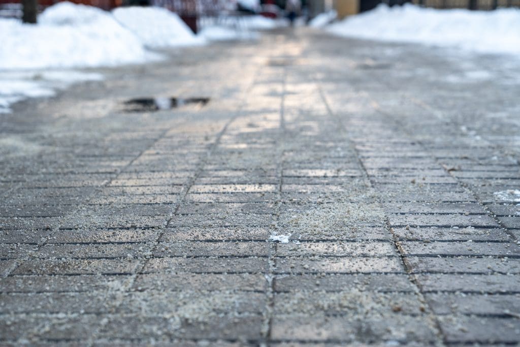 Close-up view of a brick sidewalk covered with patches of sand and ice, with snow piled along the edges and a blurry background showing more snow and distant figures.