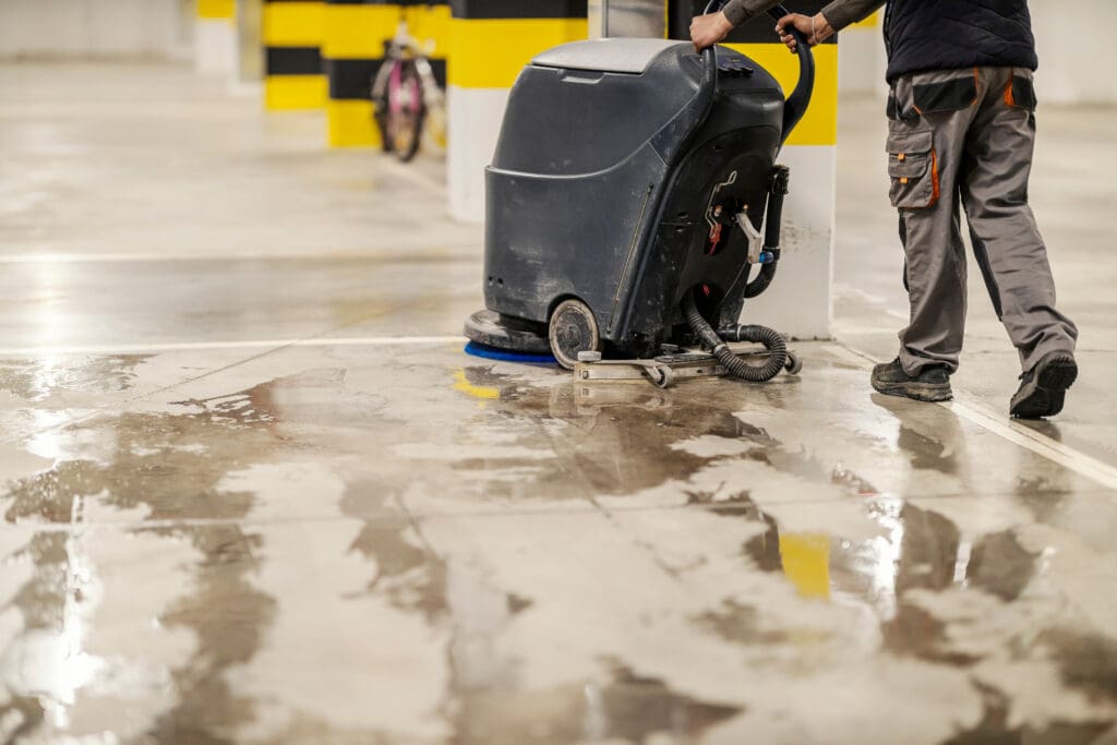 A person operates a floor cleaning machine on a wet concrete surface in a parking garage, with yellow and black pillars visible in the background.
