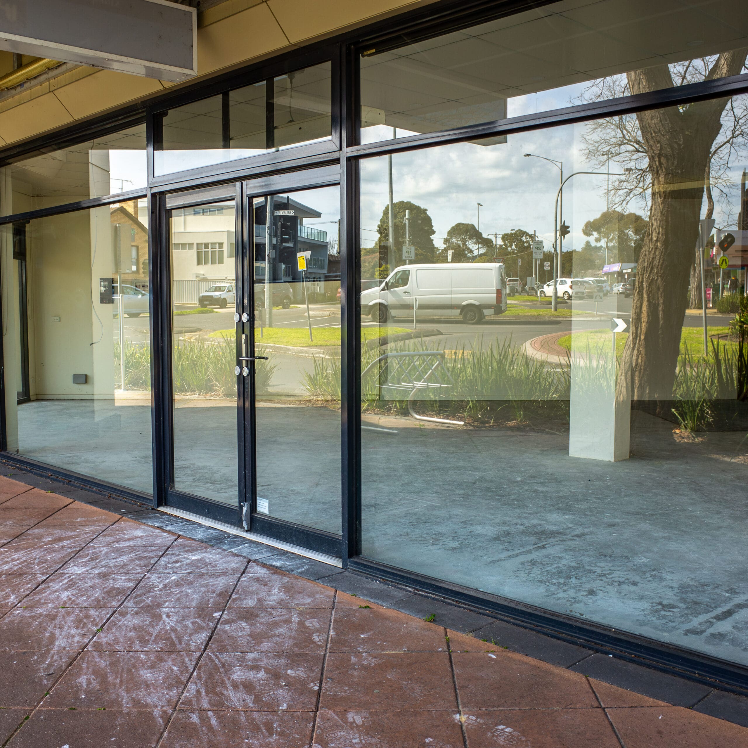 Street view of a vacant retail store with large glass windows and doors, reflecting trees, buildings, and a white van parked outside on a sunny day. The interior is empty with a bare concrete floor.