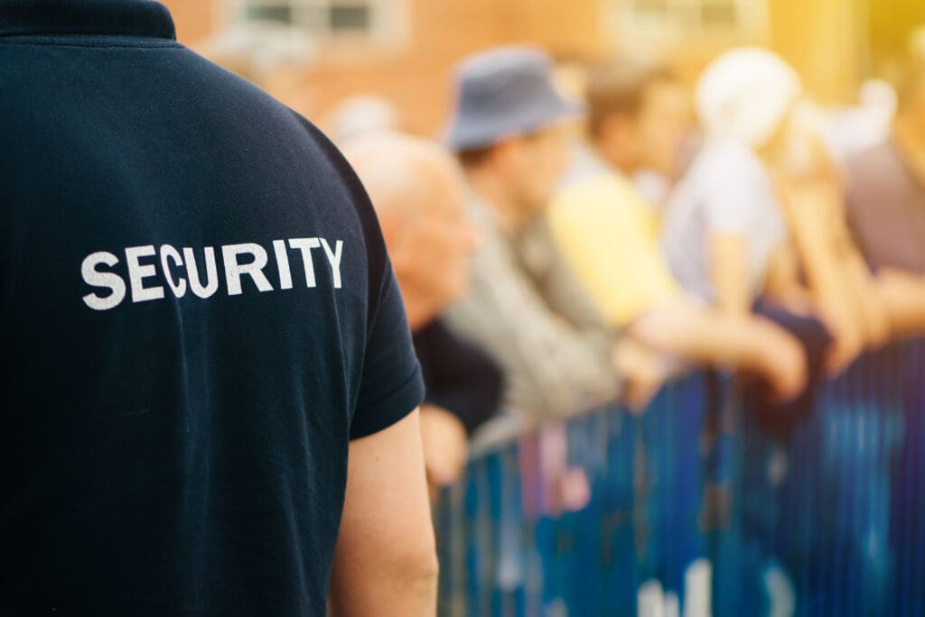 A security guard in a black shirt with "SECURITY" written on the back stands near a blue barrier, facing a blurred crowd of people at an outdoor event.