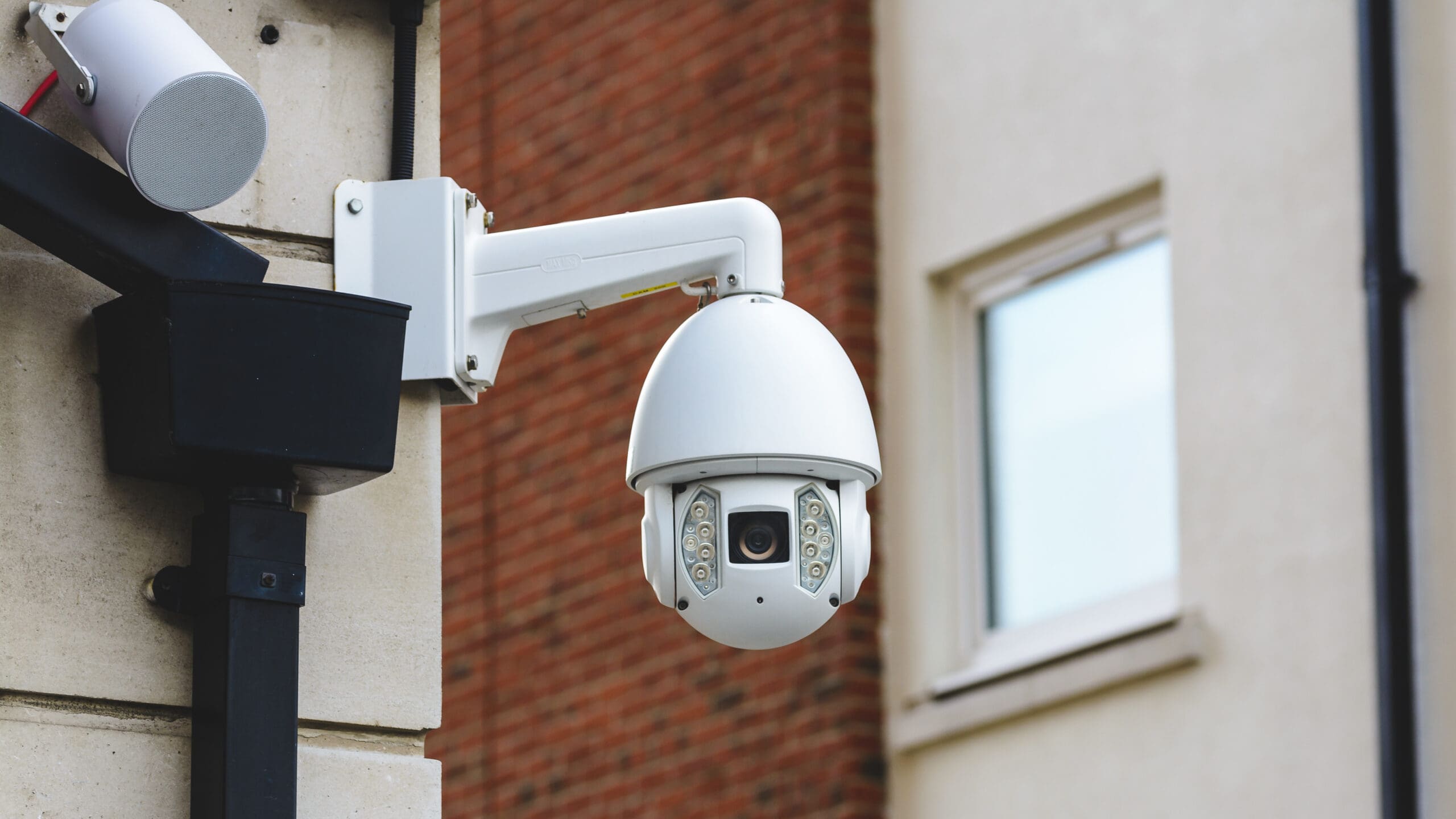 A white dome security camera is mounted on the corner of a building, with a brick wall and a window blurred in the background. The camera is angled to monitor the surrounding area.