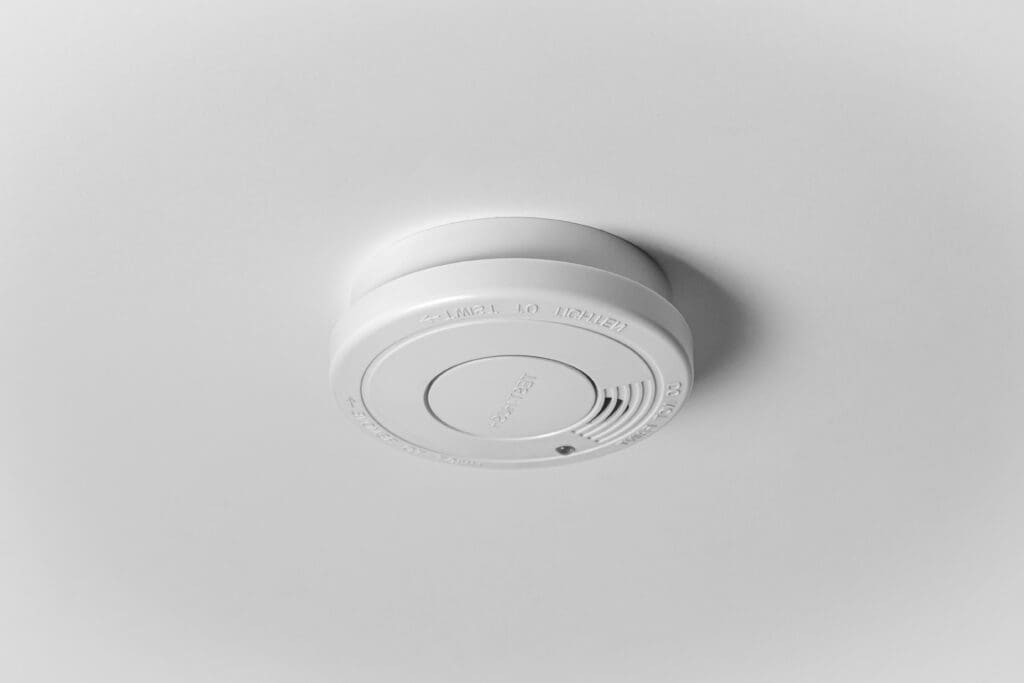 A white smoke detector mounted on a white ceiling, with a circular design and a small ventilation grille visible.