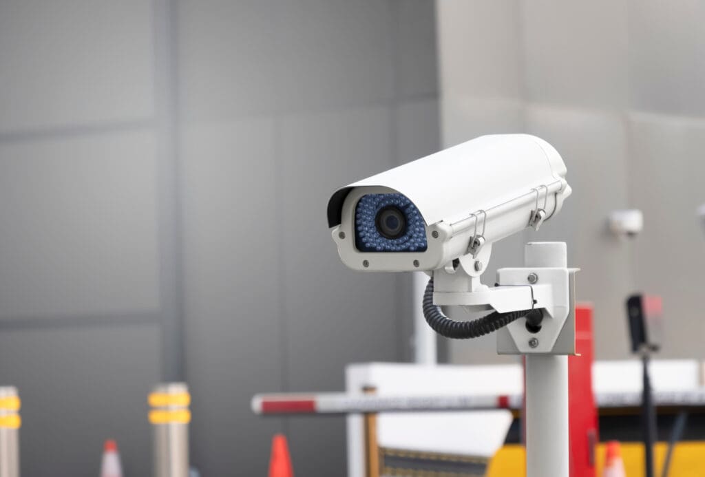 A white security camera mounted on a pole is pointed slightly upward. The background is blurred, showing barriers, an orange traffic cone, and a gray wall, suggesting a monitored area such as a parking lot or building entrance.
