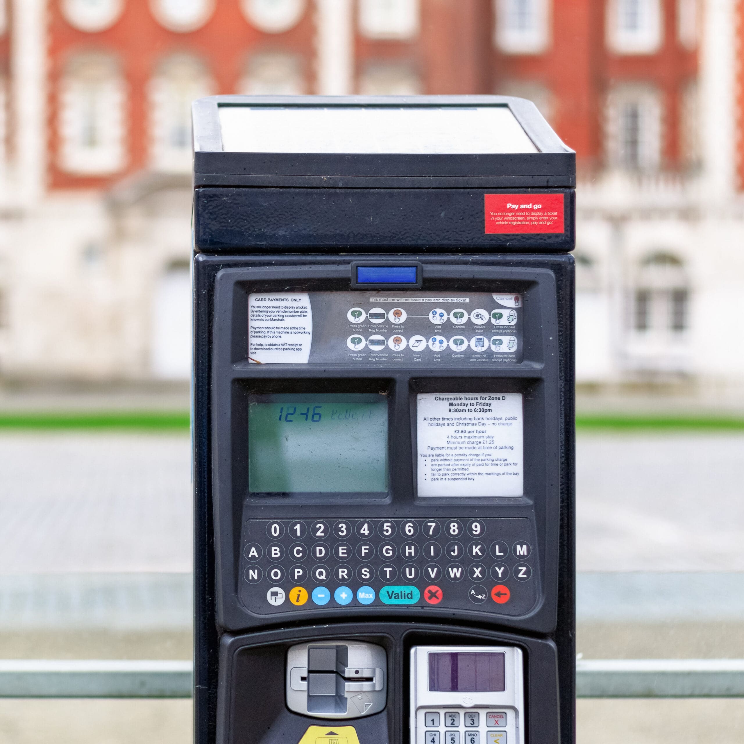 A close-up of a parking meter with buttons, a display screen showing the time, and payment slots, set outdoors with a blurred brick building in the background.
