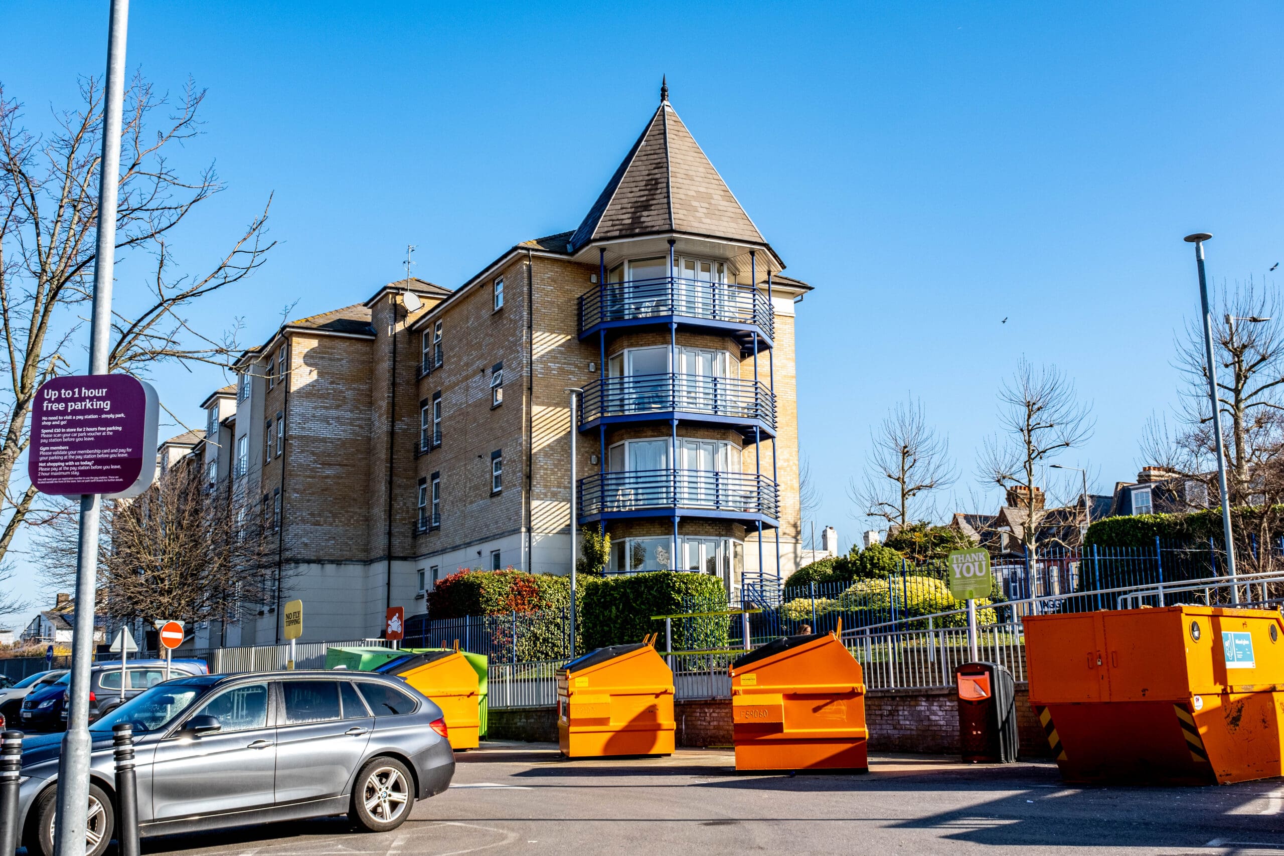 A modern residential building with a pointed roof and blue balconies stands behind a row of orange dumpsters and parked cars under a clear blue sky. A purple sign about free parking is visible on the left.