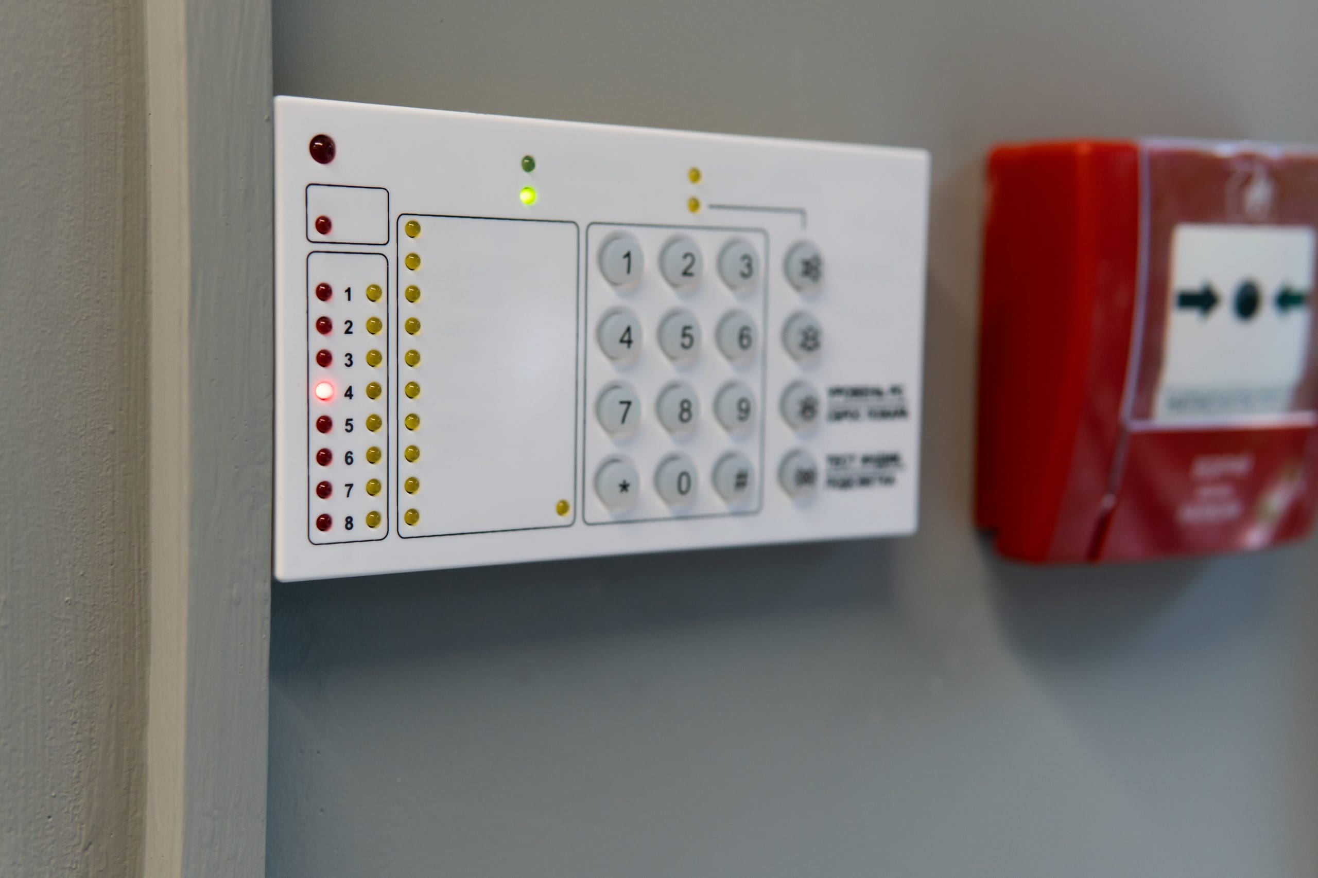 A white security alarm keypad with numbered buttons and indicator lights is mounted on a gray wall next to a red fire alarm. Several indicator lights are illuminated on the keypad.
