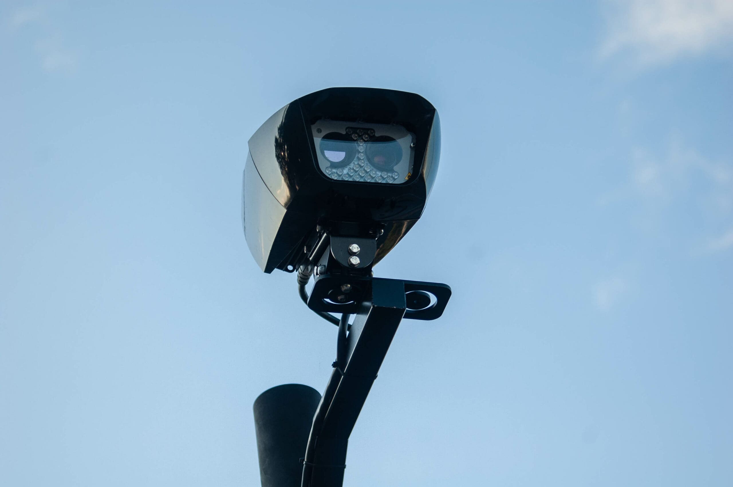 A close-up view of a black surveillance camera mounted on a metal pole, set against a clear blue sky.