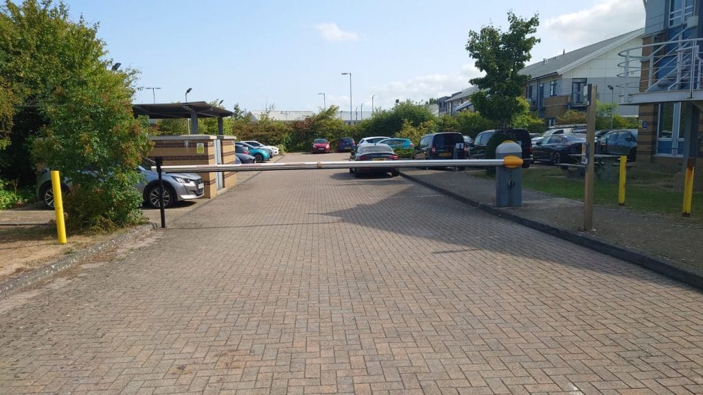 A parking lot entrance with a lowered barrier arm blocking access. Several cars are parked on both sides, and a road leads further into the lot. Trees and a building are visible in the background.