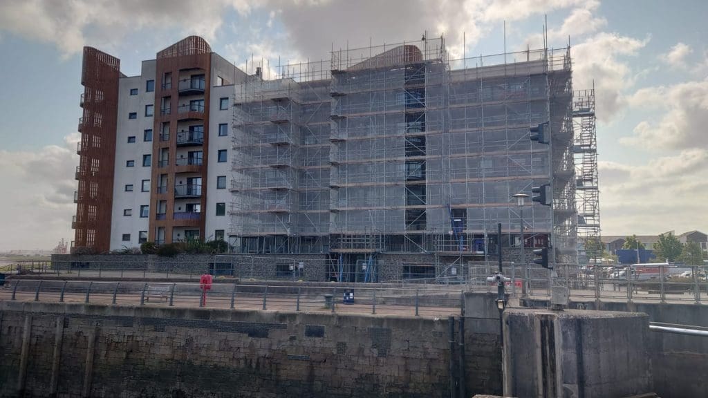A multi-storey apartment building with scaffolding on one side, set against a cloudy sky. A stone wall and metal railings are in the foreground, with a dock or canal visible at the bottom of the image.