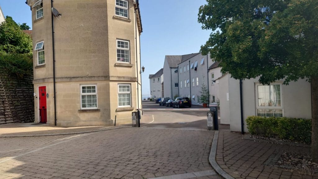 A paved street leads between stone buildings with white-trimmed windows. A red door stands on the corner building, while several parked cars are visible down the road. A large tree shades the right side.