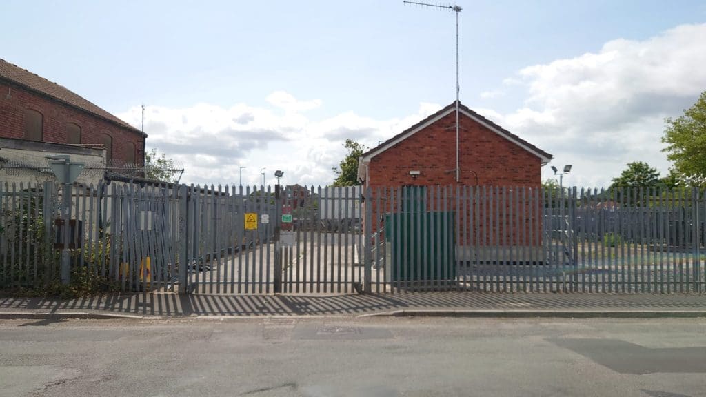 A fenced facility with a metal gate, brick building, security cameras, and warning signs. The area is empty, with blue sky and scattered clouds overhead. Trees and industrial buildings are visible in the background.