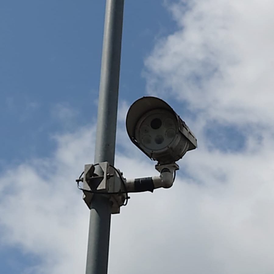 A security camera mounted on a metal pole, angled downward, with a partly cloudy blue sky in the background.