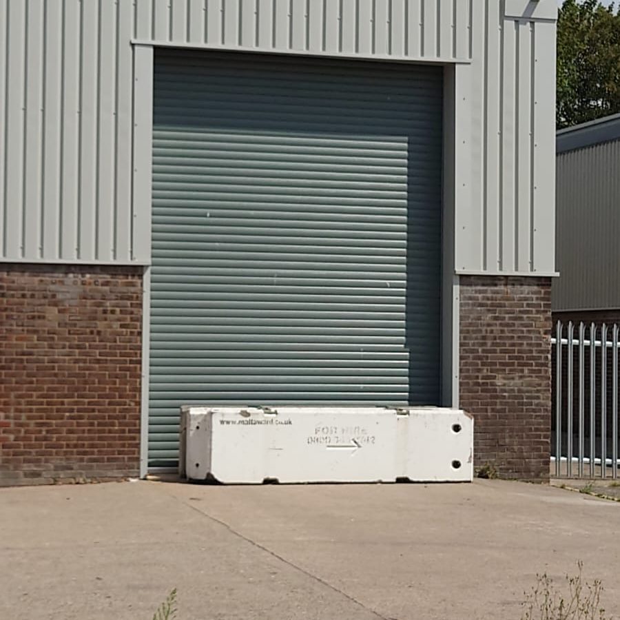 A large metal roller shutter door on an industrial building is blocked by a white concrete barrier on a concrete driveway. The building has brickwork at the base and vertical metal siding above.