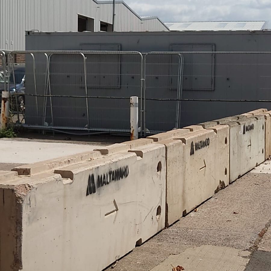 Concrete barriers labeled "MALTATMAWD" line the edge of a paved area near a construction site with metal fencing and a gray portable building in the background.