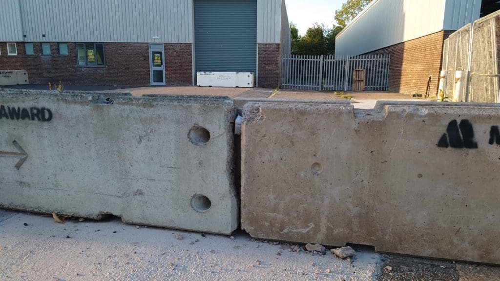 Two large concrete barriers block the entrance to an industrial building with a closed roller shutter door, metal fencing, and brick walls visible in the background.
