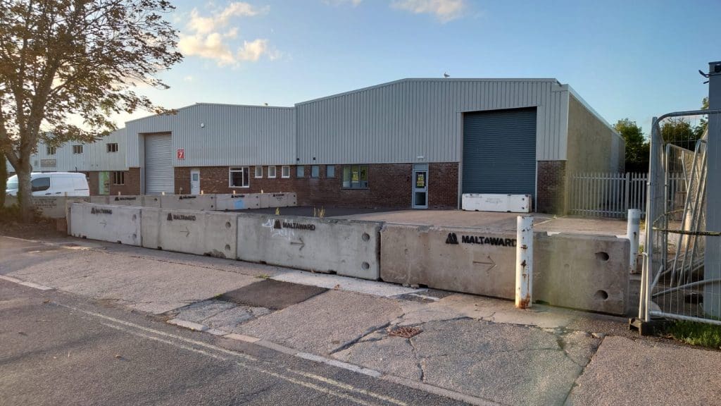 A row of large concrete barriers lines the front of a modern industrial warehouse with gray metal siding and brick base. A closed roller shutter door and a van are visible. The sky is clear with some clouds.